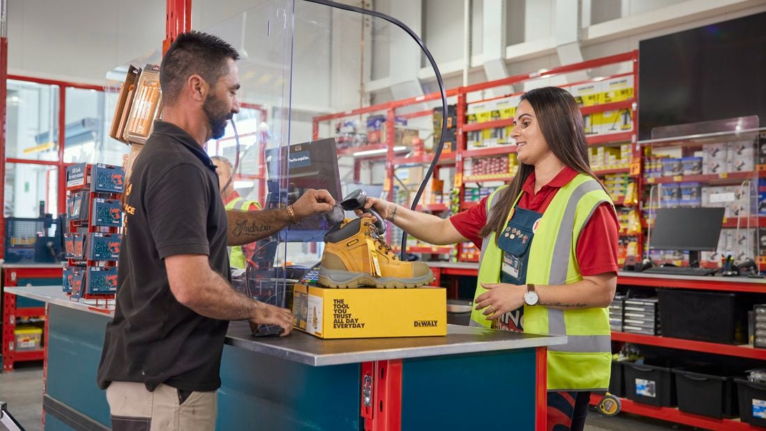A male tradie holds up his Bunnings Trade PowerPass App to be scanned by a Bunnings Warehouse Red Shirt team member in store, with yellow DeWALT boots on top of a box on the counter.