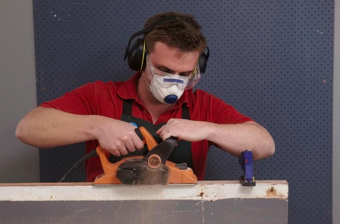 A person wearing protective gear planing the edge of a piece of timber using an electric planer