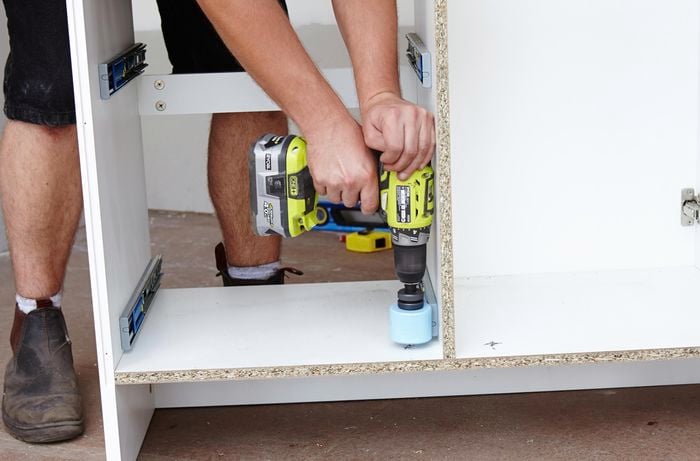 A person using a hole saw attachment to cut holes in the bottom of a bathroom vanity