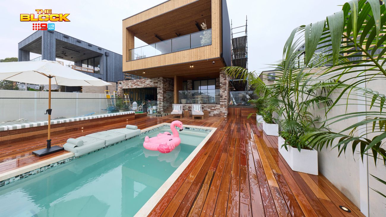 Modern outdoor pool area featuring timber decking, tropical plants, sun umbrellas and a two-storey home.