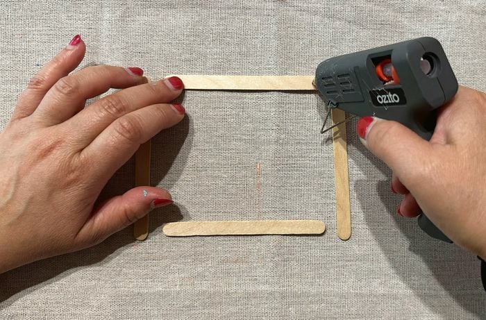 Hands assembling a square frame made of wooden craft sticks, using a hot glue gun on a fabric-covered surface.