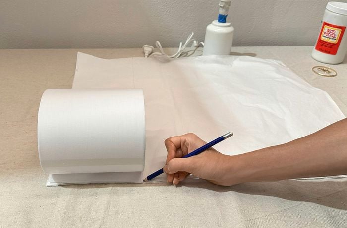 A white lamp shade on white tissue paper with a hand holding a pencil marking the height