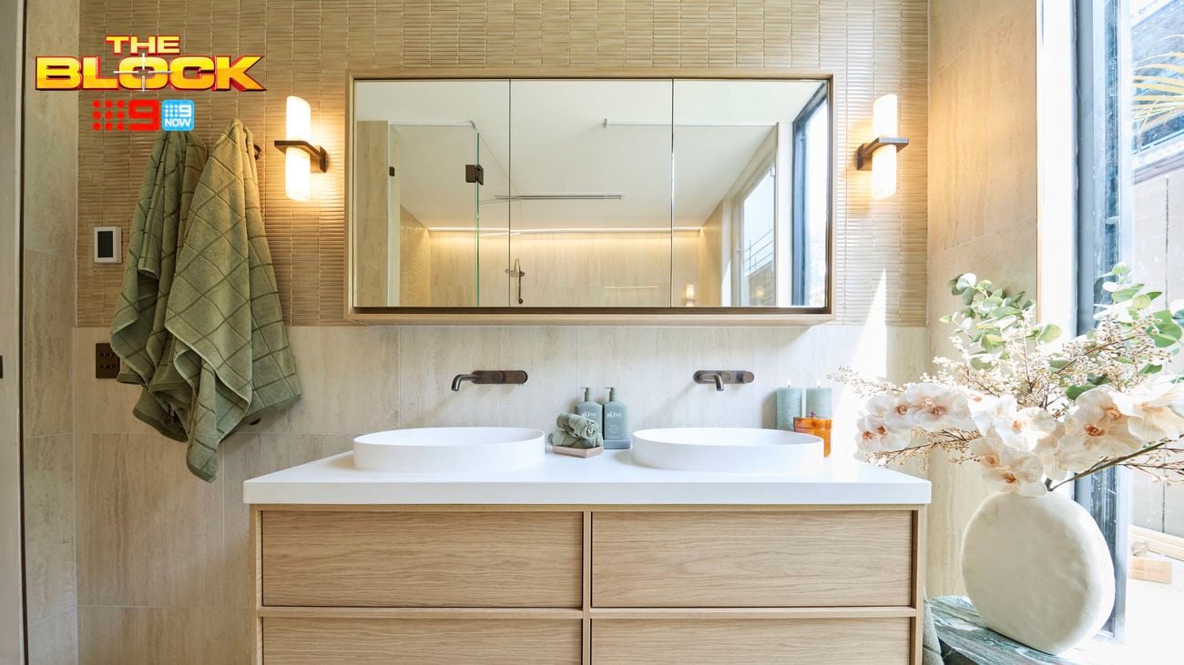 A modern bathroom with two sinks with a drawers unit under them, mirror on the wall, towels hanging on hooks on the wall, table with a vase with flowers in it on top of it.