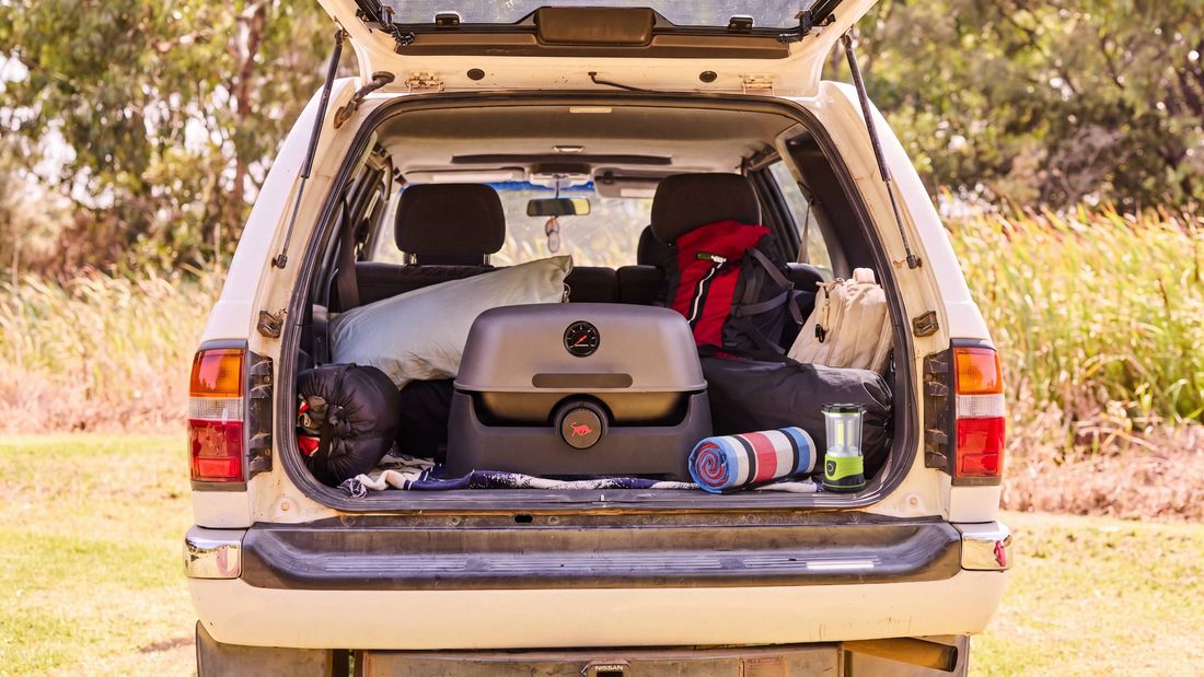 White SUV packed with camping gear and a portable BBQ, parked outdoors with the tailgate open.