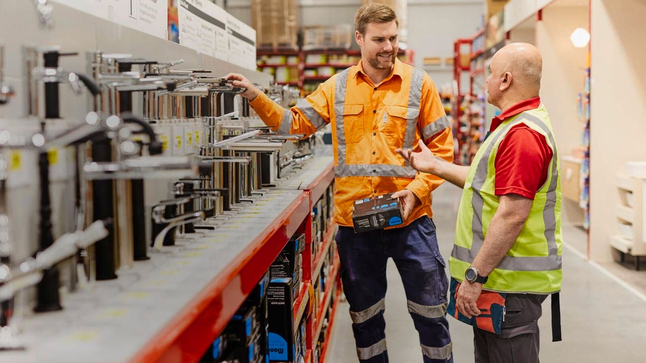 A tradie and a worker in high-visibility clothing discuss a plumbing fixture in Bunnings.