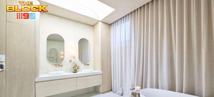 A modern bathroom with curtains, a bath, two mirrors on the wall, two sinks with drawers underneath and a wardrobe in it.