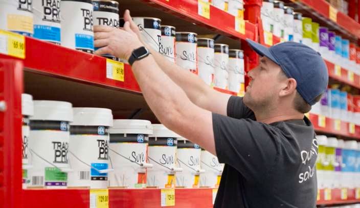 Store worker places paint buckets on a red retail shelf lined with labelled tubs and price tags in a hardware store aisle.