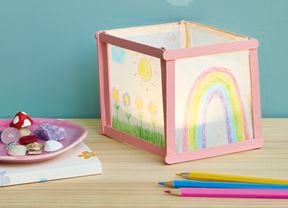 A handmade paper lantern decorated with children’s drawings of flowers, a sun, and a rainbow, sitting on a wooden table alongside coloured pencils and a small dish of shells and trinkets.
