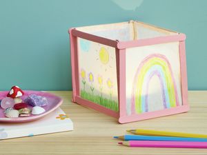 A handmade paper lantern decorated with children’s drawings of flowers, a sun, and a rainbow, sitting on a wooden table alongside coloured pencils and a small dish of shells and trinkets.