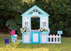 A small wooden cubby house painted white and blue, with window boxes holding white flowers, and a picket fence, sits in a garden.
