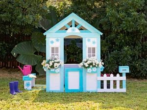 A small wooden cubby house painted white and blue, with window boxes holding white flowers, and a picket fence, sits in a garden.
