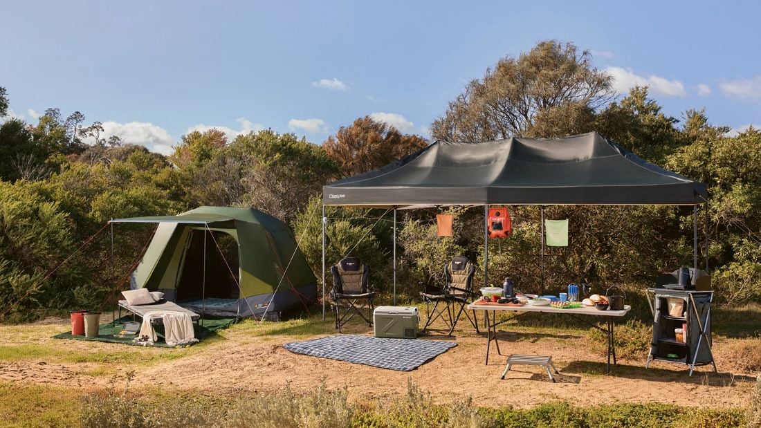 A campsite set up on grass with a green dome tent, a gazebo shelter, camping chairs, a folding table with cooking gear, and a portable stove, positioned near bushland.