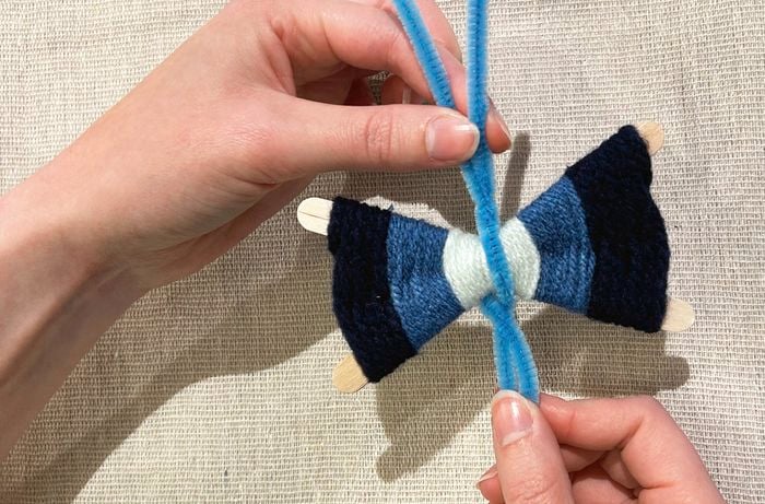 Two hands hold the end of two blue pipe cleaners across black, navy and white woven yarn on paddle pop sticks.