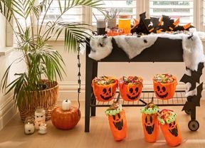 A Halloween craft display by a bay window with a skeleton figure, a decorated trolley holding treats and drinks, hanging bat-themed lanterns, and pumpkin buckets on the floor.