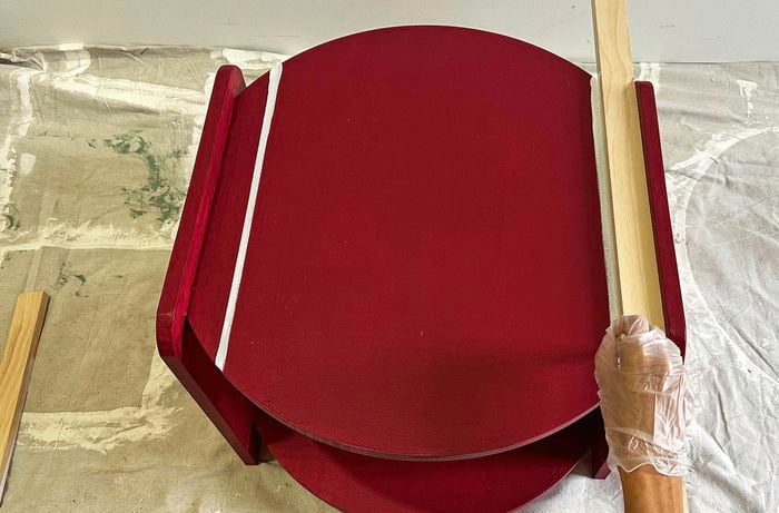 A gloved hand holds a piece of timber on a red two tier coffee table that has two white stripes sitting on a drop sheet