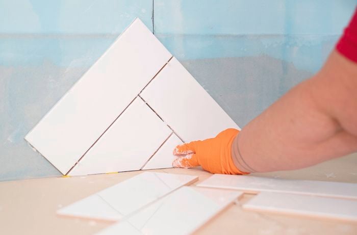A person wearing gloves is laying tiles into adhesive on a wall, forming a herringbone pattern.