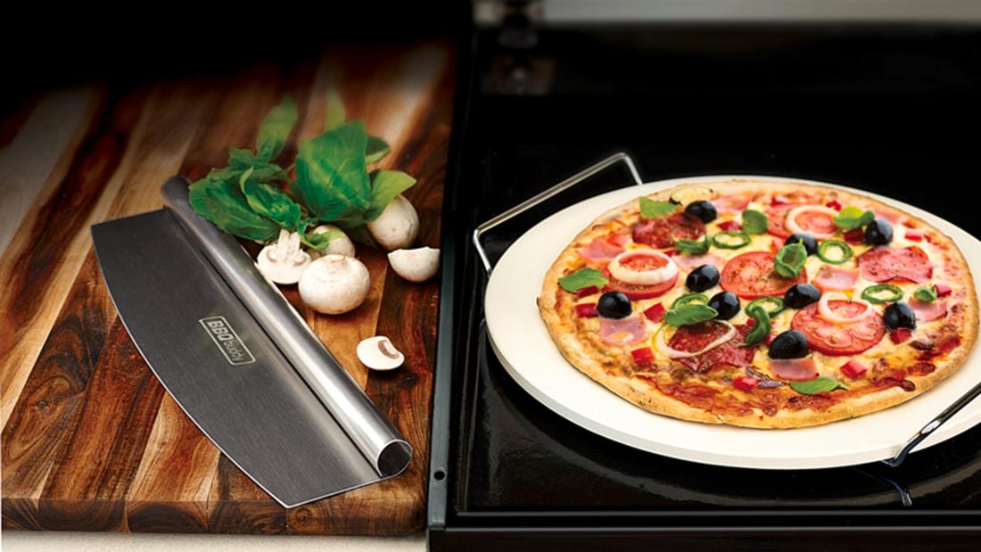 A cooked pizza on a pizza stone, with nearby cutter, herbs and button mushrooms