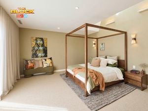 A stylish bedroom featuring a central four-poster bed with white and green bedding, multiple pillows, and a brown throw blanket. A wooden nightstand with books and decor sits to the right, while a cushioned bench with decorative pillows lines the left wall. The room includes a textured rug, light beige walls, wall-mounted lights, and artwork above the bench and bed. Sheer curtains cover large windows, allowing natural light in. 
