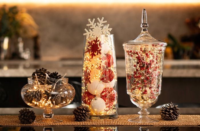 Three vases on a table are decorated with string lights, one has a pinecone, one has baubles, and one has artificial red berry stems