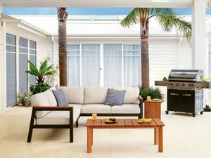 Covered outdoor entertaining area featuring a cushioned sofa, timber coffee table, barbecue and palm trees.