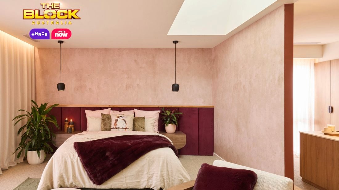 A modern bedroom with a neatly made bed featuring white and burgundy bedding, flanked by potted plants and pendant lights. Behind the bed is a light-colored wall with a wooden accent. On the right, a wooden wardrobe with open shelves stands next to an armchair with a burgundy throw pillow. A round ottoman sits at the foot of the bed, and a small table with candles is placed in front of the armchair. The ceiling includes recessed lighting and a skylight that brings in natural light.