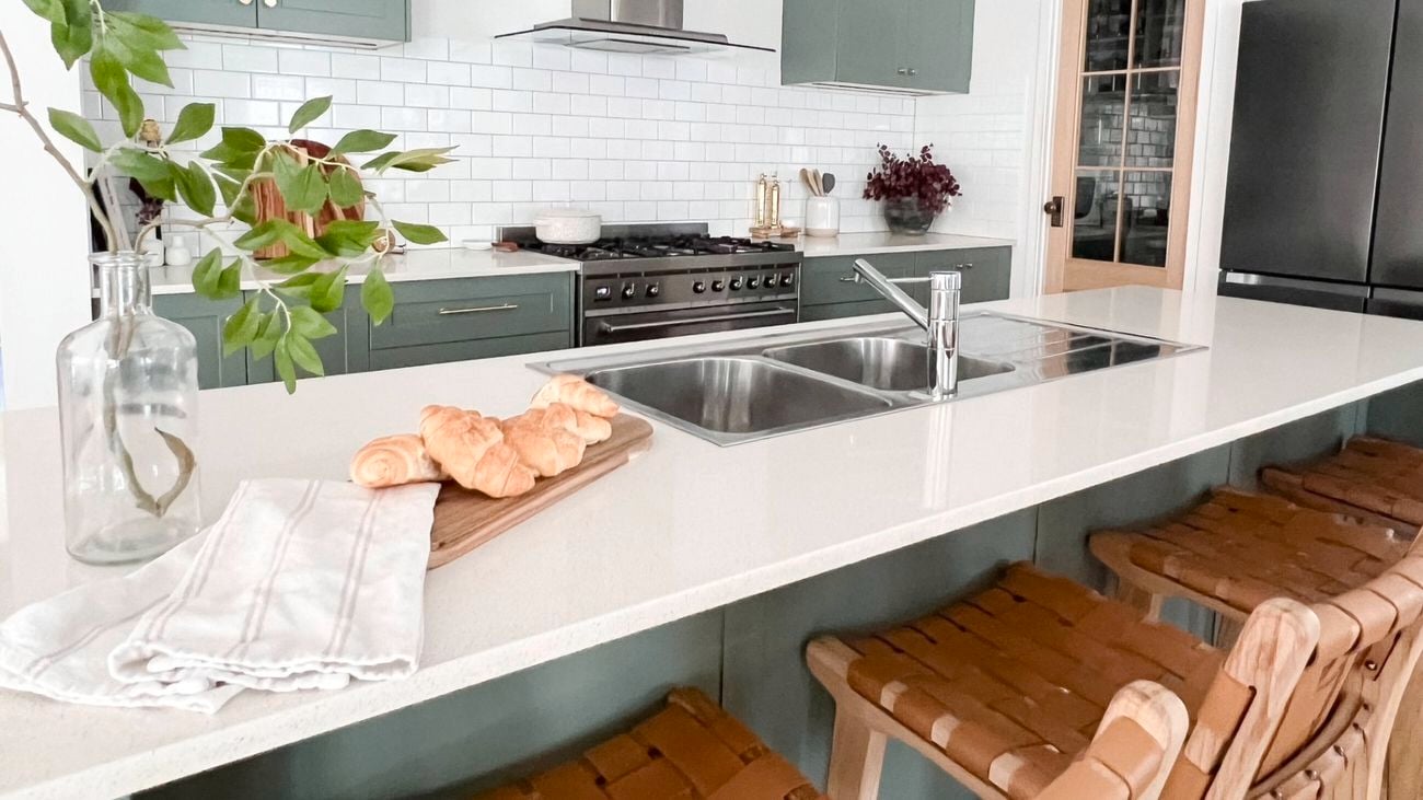 A modern furnished kitchen with a white benchtop, green cupboards and stools at the benchtop.