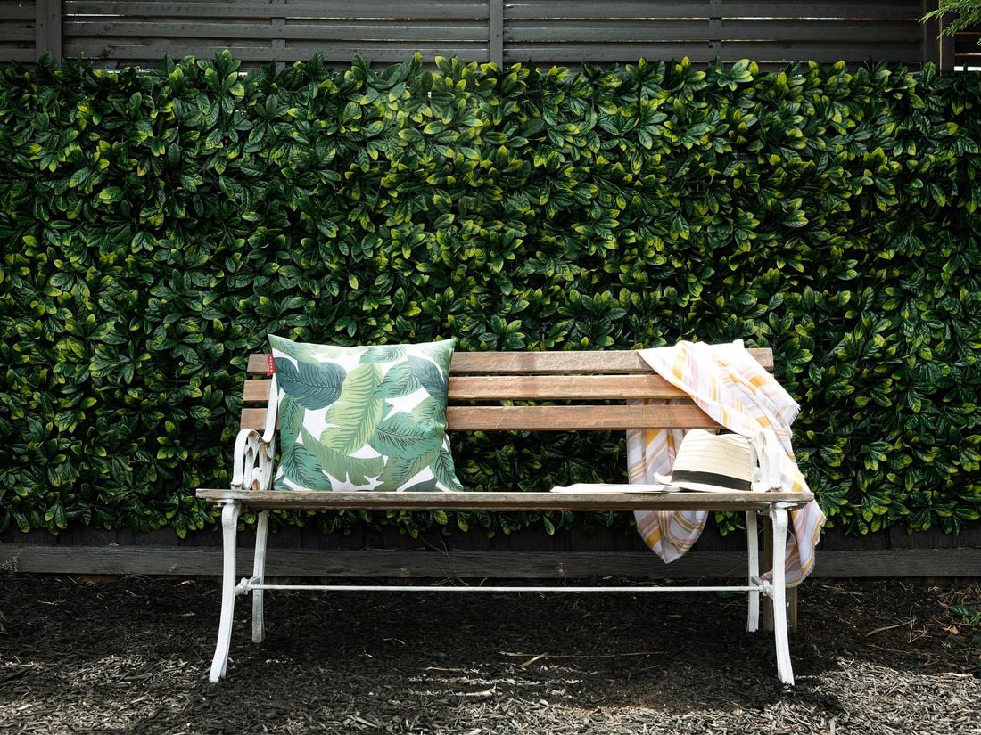 Timber benchtop in front of green hedge, with hat and blanket on it.