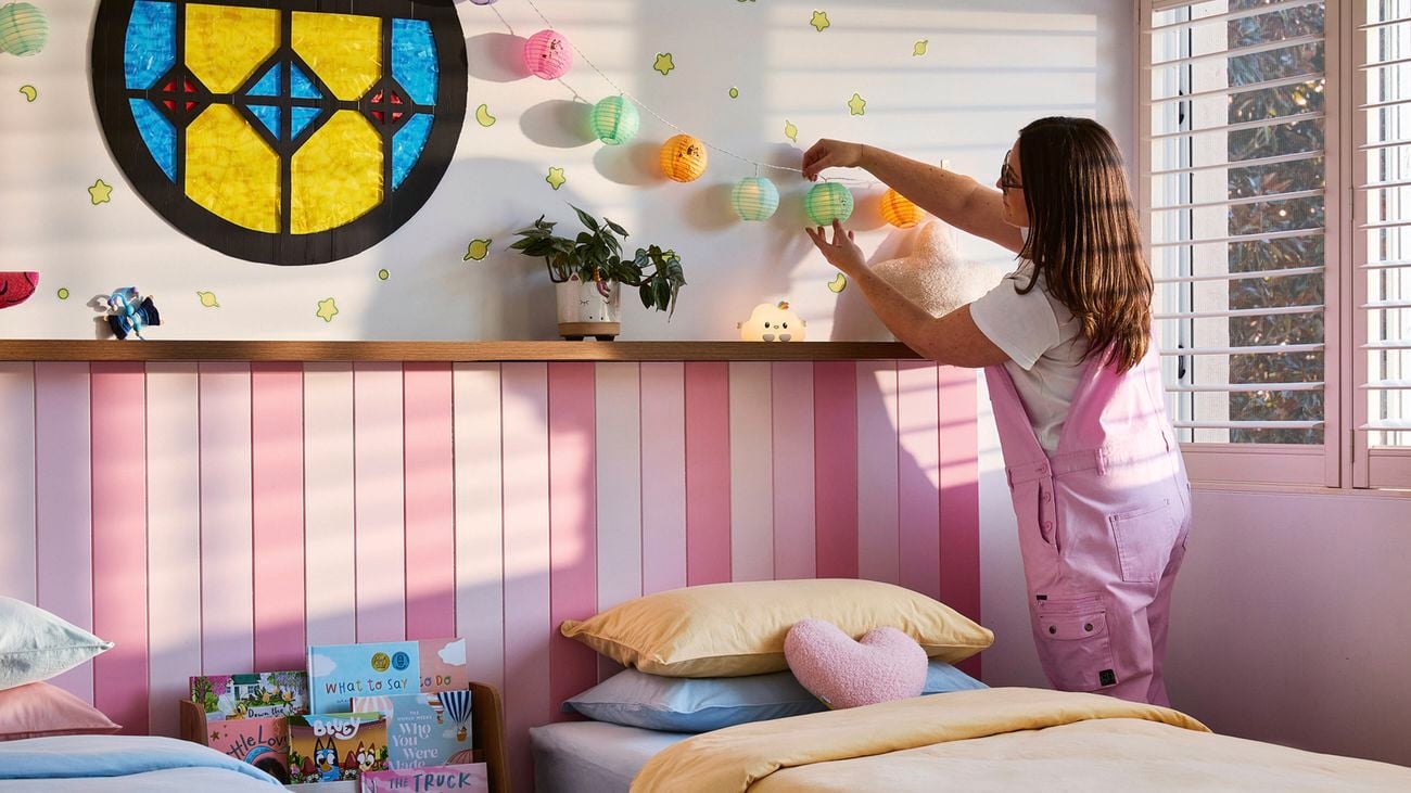 A lady with brown hair in pink overalls hangs coloured lanterns in a Bingo and Bluey bedroom, inspired by the hit TV series