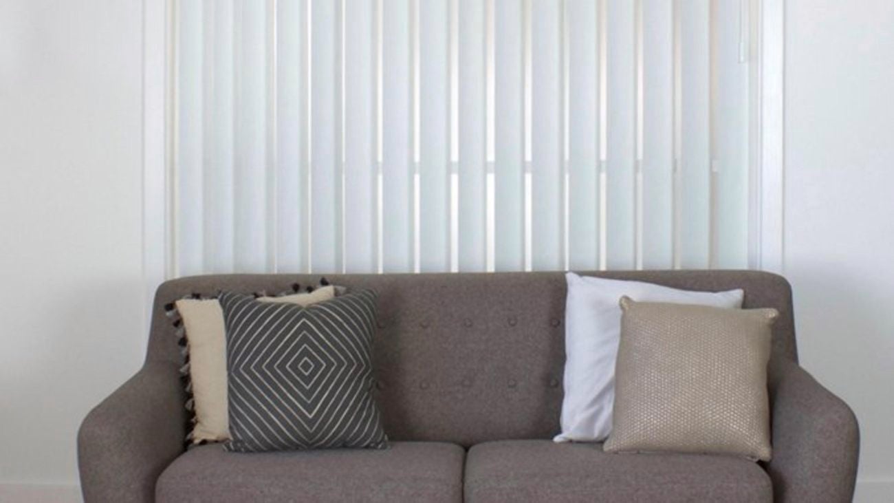 White vertical blinds behind a grey couch with cushions.