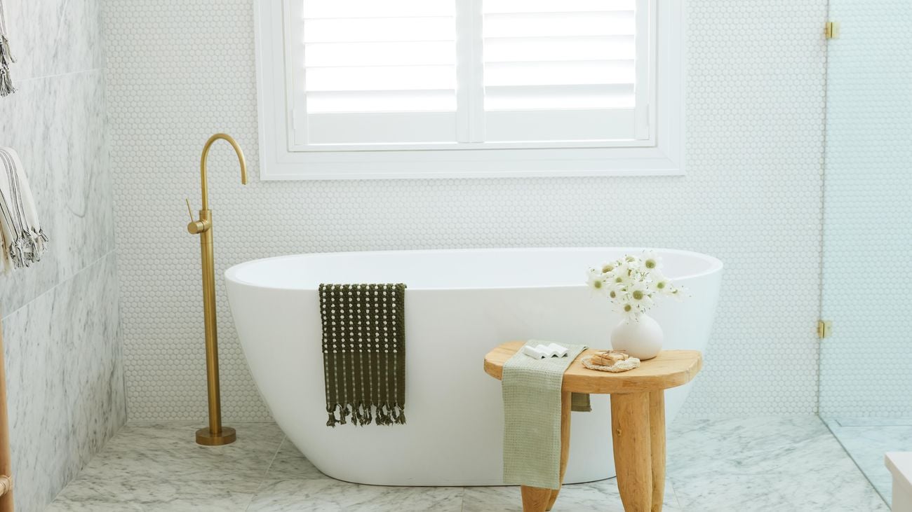 A shot of a bathroom with a white, bathtub and small wooden table, sits in front of it with a vase of flowers on top