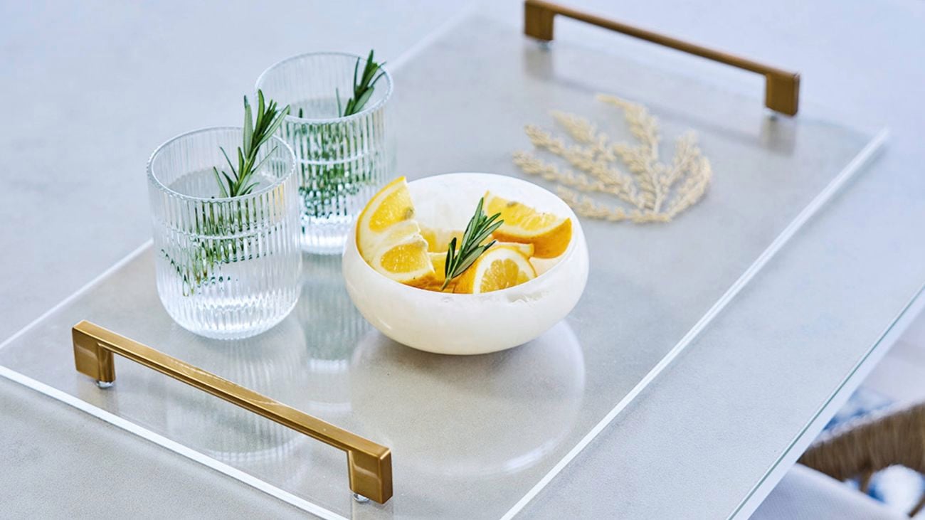   A clear rectangular serving tray with brass handles on a light-colored table, holding two ribbed glasses of water garnished with rosemary and a white bowl filled with lemon wedges, with a wicker chair and white railing in the background.