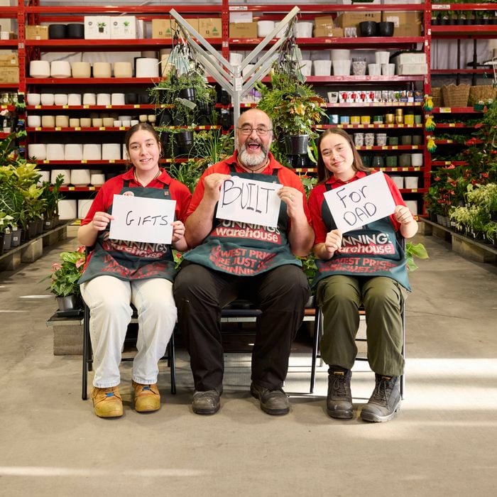 Three Bunnings team members seated in the garden centre holding signs that read “Gifts”, “Built” and “For Dad”, surrounded by indoor plants and pot displays.