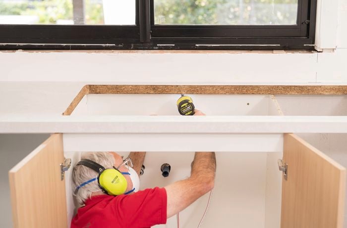 A Bunnings Team Member is is drilling upwards from inside a cabinet to fix the benchtop with screws.
