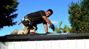 Person kneeling on a metal roof and using a cordless drill to install roofing materials outdoors, with trees and a clear blue sky in the background.