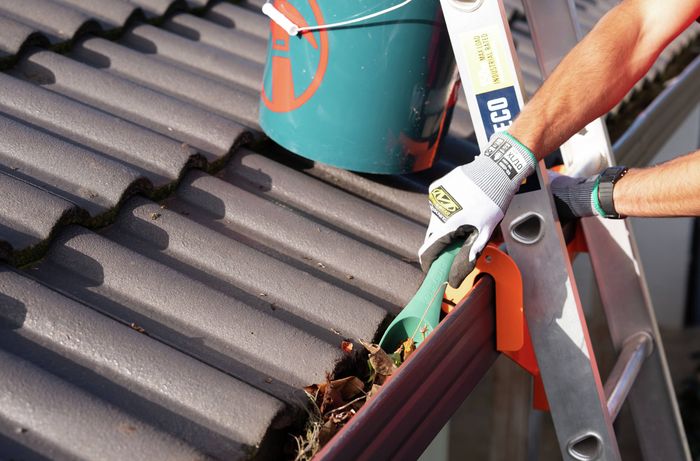 Close-up of a person using a gutter scoop to remove leaves and debris from a roof gutter while standing on a ladder.