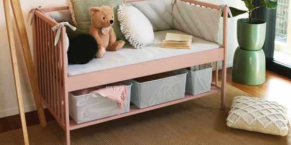 Completed light pink reading nook carrying grey pillows and an open book. 2 Grey mesh storage containers carry a blanket and books underneath the seats
