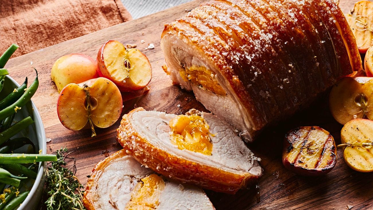 Cooked porchetta with apricot filling on a wooden chopping board, sliced, surrounded by sliced grilled apricots and assorted greens.