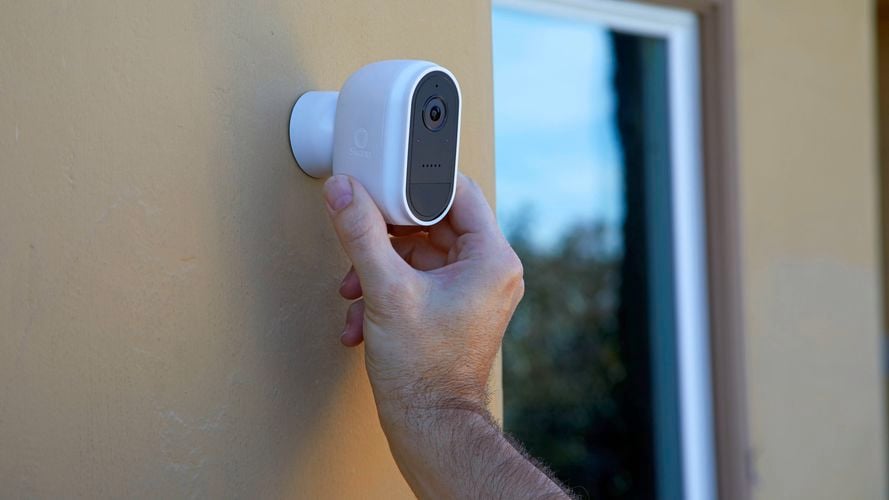 Hand adjusting a small white security camera mounted on an exterior wall near a window.
