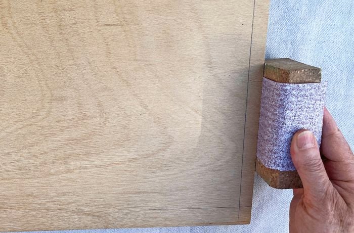 A hand sanding the edge of a piece of plywood with a sanding block