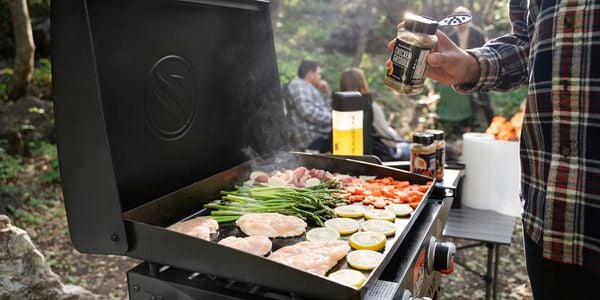 Blackstone griddle with hood and cart and a person cooking and seasoning chicken and vegetables.