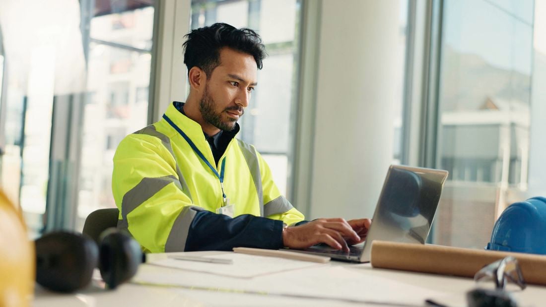 A man in a high-vis vest works on a laptop at a desk with construction plans and a hard hat