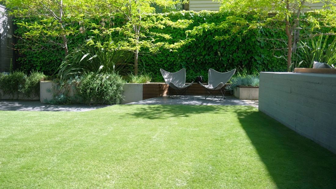 A large backyard with a smooth green lawn of Tuff Turf artificial grass and paved seating area plus raised garden beds.