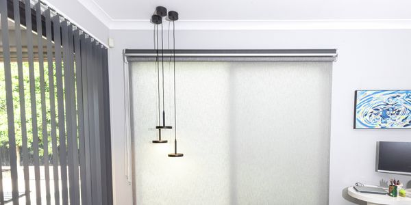 Light grey roller blind covering a large window in a modern room, with dark vertical blinds on a sliding glass door to the left and black pendant lights hanging in front of the window.