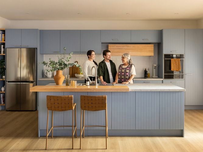 Couple in their 30's and their Nonna are standing at an island bench in a light blue kitchen. They are smiling at each other.