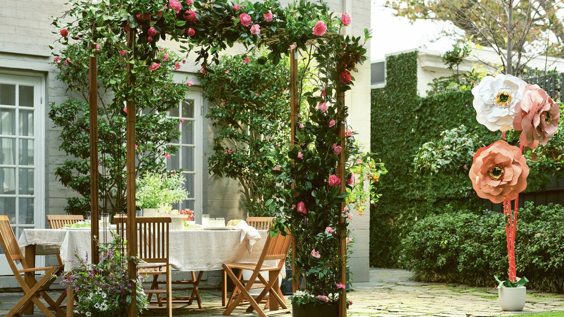 Outdoor garden arch with pink flowers and foliage in front of a dining table, with a paved patio and grassy lawn