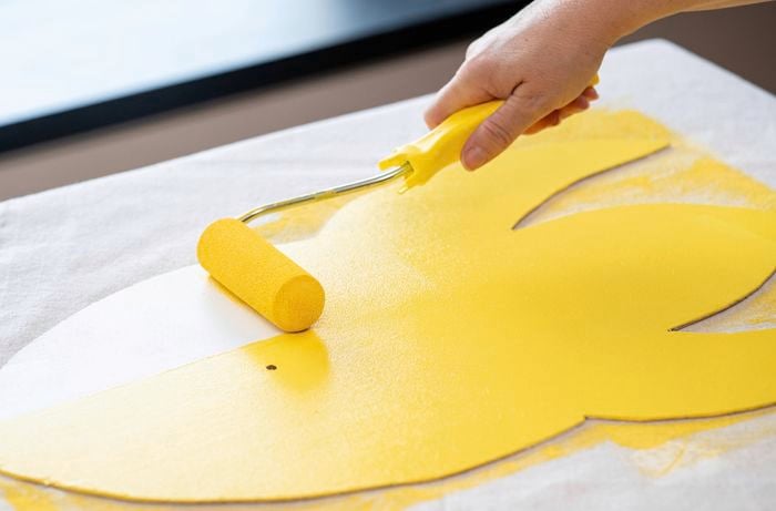 A person is applying bright yellow paint to a plywood rocket shape with a paint roller.