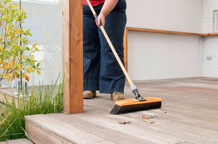 A person is sweeping a wooden deck with a broom featuring an orange and black brush.