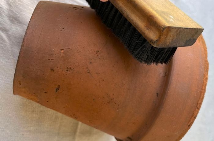 A dirty terracotta pot being scrubbed with a firm-bristled brush on top of a drop sheet.