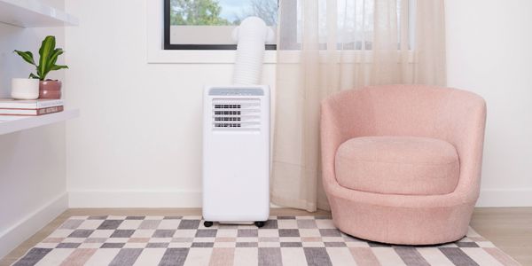 Portable air conditioner with window exhaust hose installed beside a pink armchair in a bright room with sheer curtains and a patterned rug.