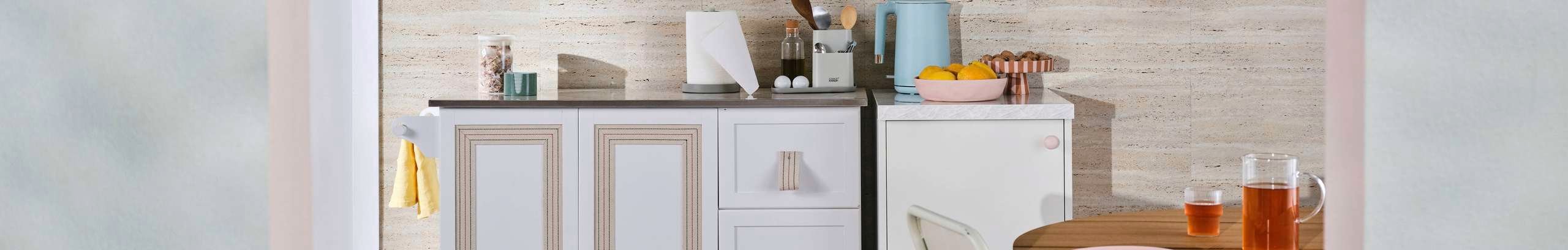 A styled compact kitchen with white cabinetry, pastel accents and a wooden dining table, set against a neutral stone-look splashback.